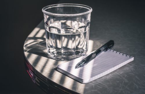 Ein Glas Wasser neben Notizblock und Stift auf Tisch.
